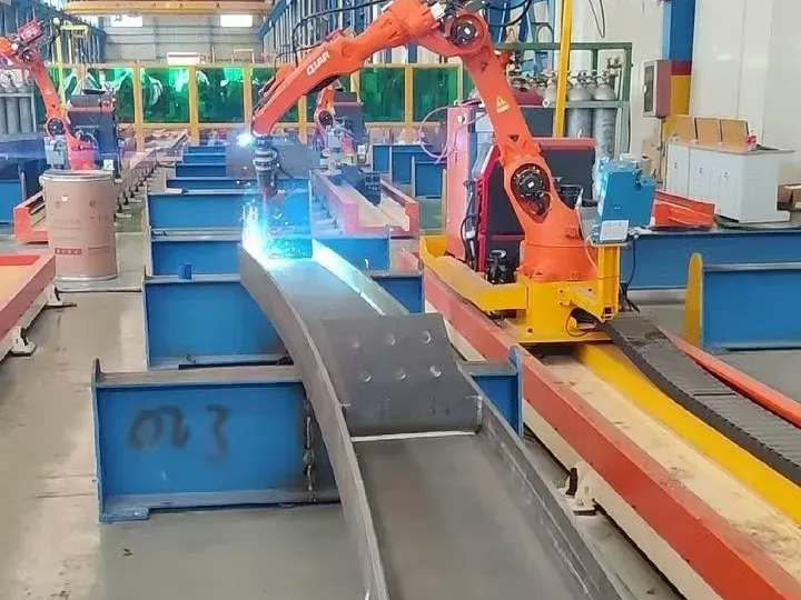 Orange rail-mounted robot welding a curved steel beam, with more stations behind.