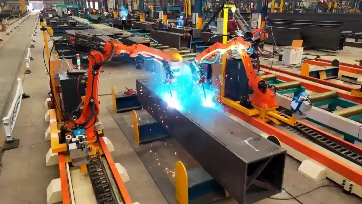 Two orange rail-mounted robots welding a steel box beam, with sparks at both torches.