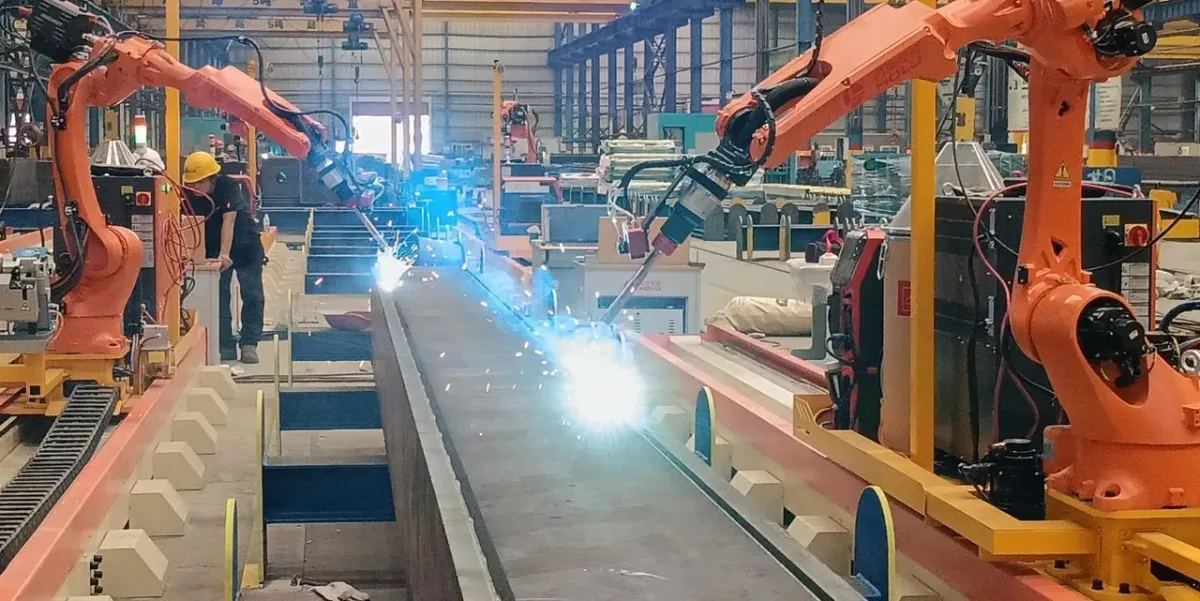 Two orange welding robots on ground rails welding a steel member, with bright arcs and sparks.
